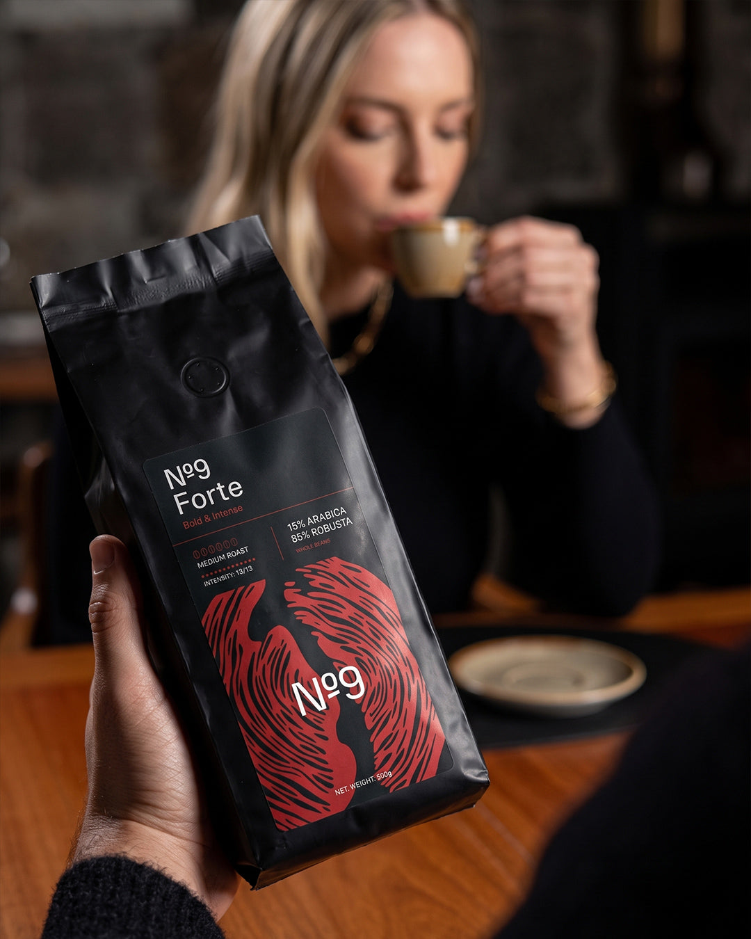 Vrečka kave v zrnju No.9 Caffè Forte v ospredju, medtem ko ženska v ozadju pije kavo.