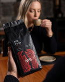 Vrečka kave v zrnju No.9 Caffè Forte v ospredju, medtem ko ženska v ozadju pije kavo.