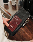 Moški drži 500 g vrečko kave v zrnju No.9 Caffè Aroma ob kavnem aparatu.